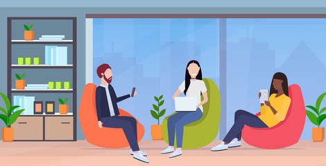 businesspeople sitting in bean bag armchair mix race colleagues team using digital gadgets social media communication concept modern office interior full length horizontal