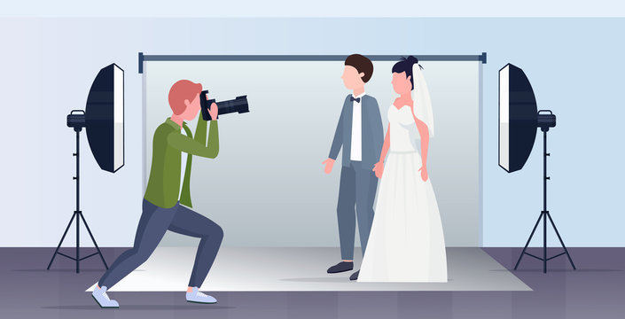 professional wedding photographer shooting on camera newly weds couple bride and groom embracing posing in modern photo studio interior full length flat horizontal