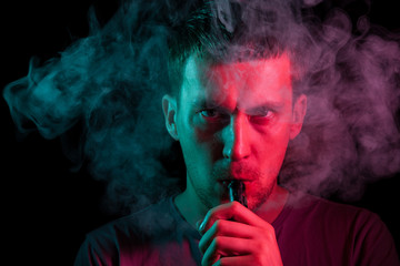 A man holds in his hand and drags on an electronic cigarette with a vape while smoking and releases smoke to the sides, his face is highlighted with red and green light, and head is shrouded in clouds