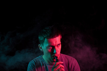 A young brunette man holds in his hand and drags on an electronic cigarette with a vape while smoking and releases smoke to the sides, his face is highlighted with red and green light.