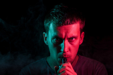 A young brunette man holds in his hand and drags on an electronic cigarette with a vape while...