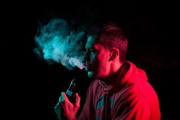 Close up portrait of the face of an adult serious man exhales green toxic smoke while smoking e-cigarette and vape illuminated with blue and red colored light on a black background. Harm to health.