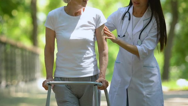 Female doctor helping to mature lady moving with walking frame, physiotherapy