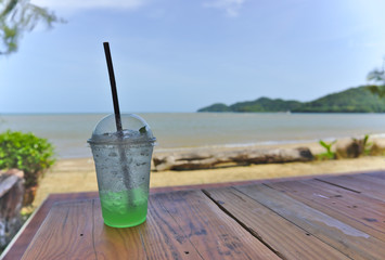 Cold iced green kiwi soda in plastic cup with brown straw on wooden table nearby white sand beach and sea