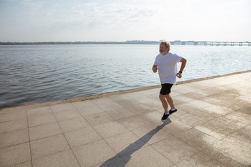 Senior man as runner with armband or fitness tracker at the riverside. Caucasian male model practicing jogging and cardio trainings in summer's morning. Healthy lifestyle, sport, activity concept.