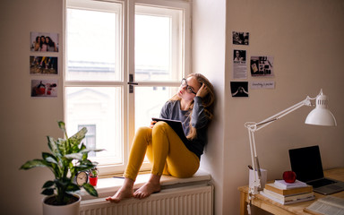 A young female student with an exercise book sitting on window sill, studying.