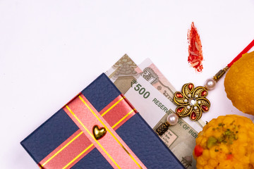 Raksha Bandhan Festival – Close view of elegant Rakhi, sweets, blue gift box, and Indian currency notes with kumkum and rice grain on a white background