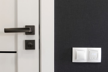 Naklejka premium White wood door with metal handle and white lighting switches on dark gray textured wall at modern apartment, copy space. Interior detail
