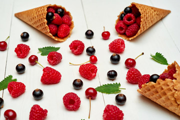 Ripe sweet raspberries, cherries and black currant in ice cream waffle cones, copy space. Fresh berries on white wood background. Summer and healthy food concept