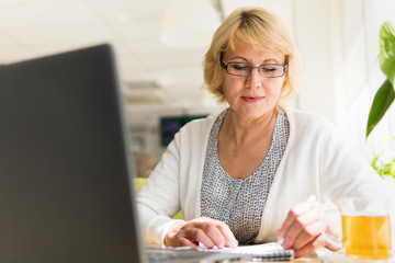 A middle-aged woman with a laptop works in a cafe in the office, she is a freelancer. A woman with glasses sits at a table with a Cup of herbal tea. She writes in a notebook.