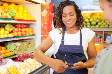 Close-up of African saleswoman in apron holding mobile phone and making a purchase with credit card in the supermarket