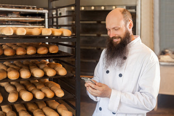 Portrait of satisfied young adult businessman baker with long beard in white uniform standing in bakery and have online order by phone, using phone with toothy smile Indoor, profession concept