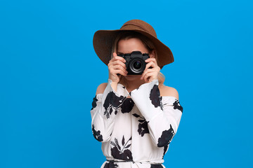 Portrait of a model with curly blond hair in a sundown hat and white dress takes photos with retro camera standing on a blue background.