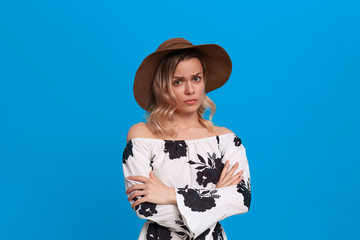 Grumpy young model with curly blond hair in a sundown hat and white dress looks at the camera...