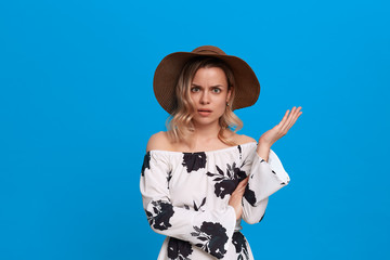 Grumpy young model with curly blond hair in a sundown hat and white dress looks at the camera...