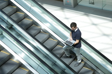 A young businessman is riding an escalator with an open laptop in his hands in the business center...