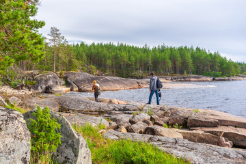 Middle aged man and young lady walking on northern lake shore in summer day. Tourists against picturesque landscape. Travelling and discovering distant places of Earth. Onega lake, Karelia, Russia