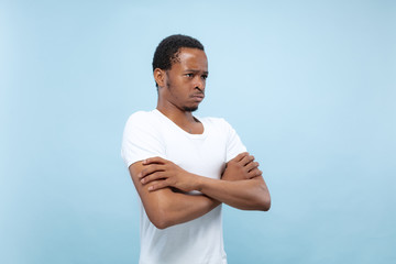 Half-length close up portrait of young african-american man in white shirt on blue background. Human emotions, facial expression, ad, sales, concept. Standing hands crossed and sad, upset or calm.