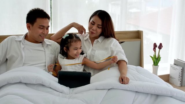 Asian family happy on the bed in the house with sunlight from the window. Father, mother and daughter, aged three, watching online media with a digital tablet. Family leisure activities, education