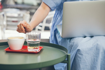 Close-up photo of a young woman dressed in casual blue dress working with laptop and drinking specialty coffee in a modern cafe with minimal Scandinavian style cafe. Selective focus, lifestyle