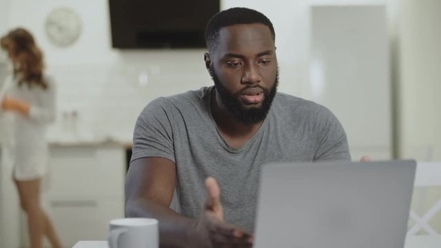 Sad black man working laptop at open kitchen. Crazy young guy typing notebook