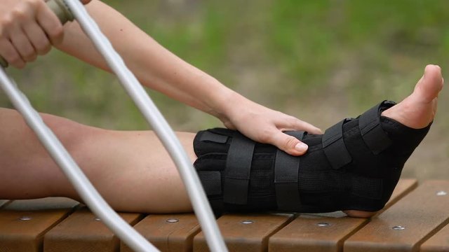 Female patient with crutches massaging painful leg in walking brace, outdoors