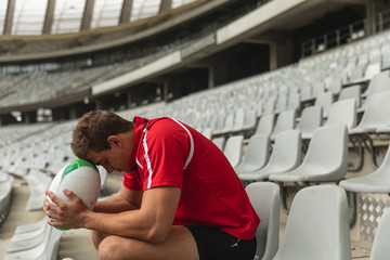 Upset Caucasian male rugby player sitting with rugby ball in stadium