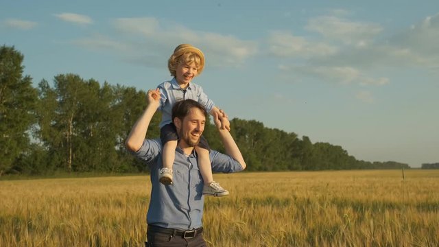 Happy father holding his little son on shoulders and walking at meadow on sunny summer day. Silhouettes of dad and his child playing at lawn. Family spending time at nature. Close up Slow motion. 4k