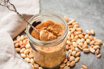 Peanut paste in an open jar and and spoon, peanuts on the gray table.