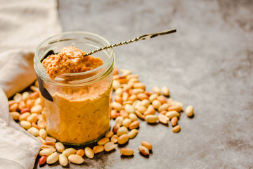 Peanut paste in an open jar and and spoon, peanuts on the gray table.Copyspace.
