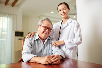 Portrait of young nurse in white coat standing near the senior man in eyeglasses who sits at the table and they smiling at camera