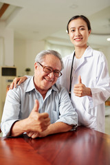 Portrait of Asian senior man and female young doctor smiling at camera and showing thumbs up together at the table