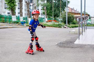 A small child in bright clothes, in a red helmet and protection goes on the road on rollers. Children's sport, active leisure.