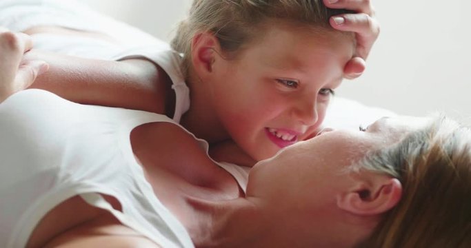 Authentic slow motion close up shot of cute smiling little girl daughter and young mother are having fun to stay together after waking up in a bed with white sheet in the morning.