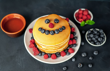 American pancakes with raspberries, fresh blueberries and honey. Healthy breakfast on concrete background, funny face on pancakes painted with blueberries