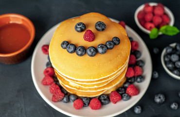 American pancakes with raspberries, fresh blueberries and honey. Healthy breakfast on concrete background, funny face on pancakes painted with blueberries