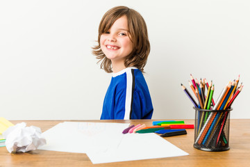 Little boy painting and doing homeworks on his desk looks aside smiling, cheerful and pleasant.