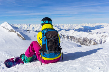 Fototapeta na wymiar Snowboarder sits on a slope with her back and looks at the panorama of the mountains.