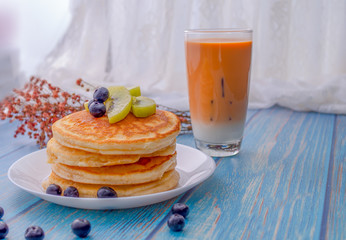 Stack of pancakes with topping, kiwi and blueberry.placed in a white plate on a blue wooden table.Eat with milk tea in the glass.