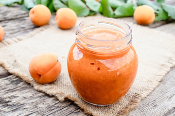 A glass jar with homemade apricot jam on a wooden surface near ripe apricot and green leaves.