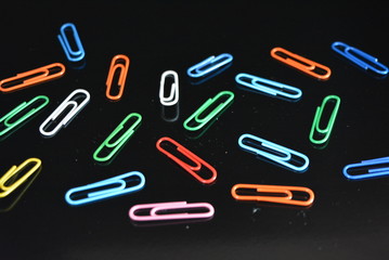 Many bright glossy colorful paper clips lie on a black glossy surface with reflection.