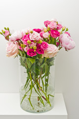 Bouquet of pink roses, peony flowers in glass vase on a shelf against white wall background.