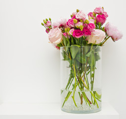 Bouquet of pink roses, peony flowers in glass vase on a shelf against white wall background.