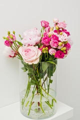 Bouquet of pink roses, peony flowers in glass vase on a shelf against white wall background.