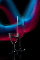 A two wine glass isolated on a black background with red, white, pink and orange neon light painting streaks of light behind them. Light wave, play of color, drawing with light.