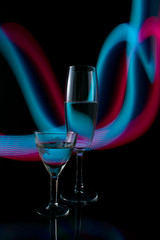 A two wine glass isolated on a black background with red, white, pink and orange neon light painting streaks of light behind them. Light wave, play of color, drawing with light.