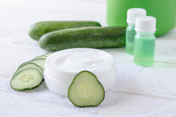 Face cream in a white jar with cucumber extract next to slices of fresh cucumbers on a white wooden background.