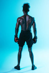 Fototapeta na wymiar back view of shirtless, muscular african american sportsman in boxing gloves on blue background