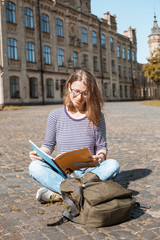 Smiling girl student reads a book while sitting near the university. A young woman is looking into a textbook while sitting near college.