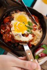 Woman is having breakfast on summer terrace: israeli shakshuka with tomatoes, salami, chili pepper and fried eggs, served in hot cast pan with toasts
